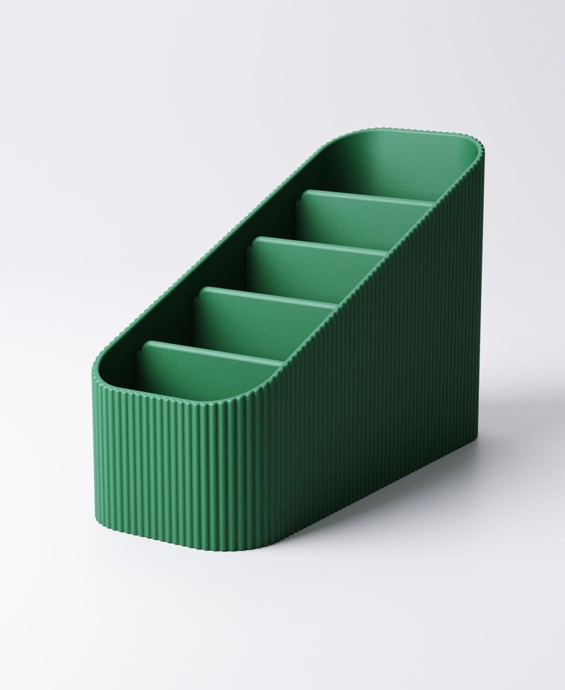 May include: A green desk organizer with a ribbed exterior and four interior compartments. The organizer has a slanted design, with the compartments increasing in height. The color is a deep, saturated green.