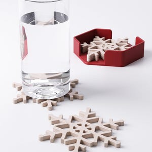May include: A clear glass of water sits next to a red holder containing several beige snowflake-shaped coasters. Two additional snowflake coasters are on the white surface. The coasters are designed to protect surfaces from water rings.