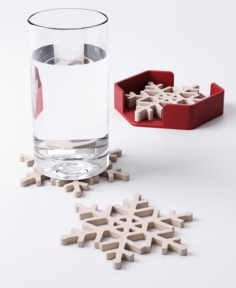 May include: A clear glass of water sits next to a red holder containing several beige snowflake-shaped coasters. Two additional snowflake coasters are on the white surface. The coasters are designed to protect surfaces from water rings.
