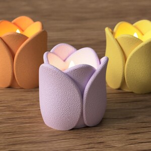 May include: Three tulip-shaped candle holders in orange, lavender, and yellow. Each holder has a lit candle inside, casting a warm glow. The holders are made of a textured material and sit on a wooden surface.