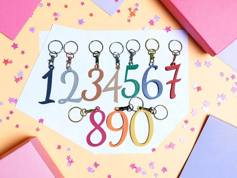 May include: A collection of keychains featuring the numbers 0 through 9 in various colors. Each number is attached to a metal keyring. The background is a light orange color with pink and purple star-shaped confetti and pink and purple gift boxes.