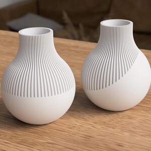 May include: Two white ceramic vases with a textured, ribbed design on the upper portion and a smooth, rounded base. One vase has a diagonal design. The vases are displayed on a wooden surface, showcasing a modern, minimalist aesthetic.