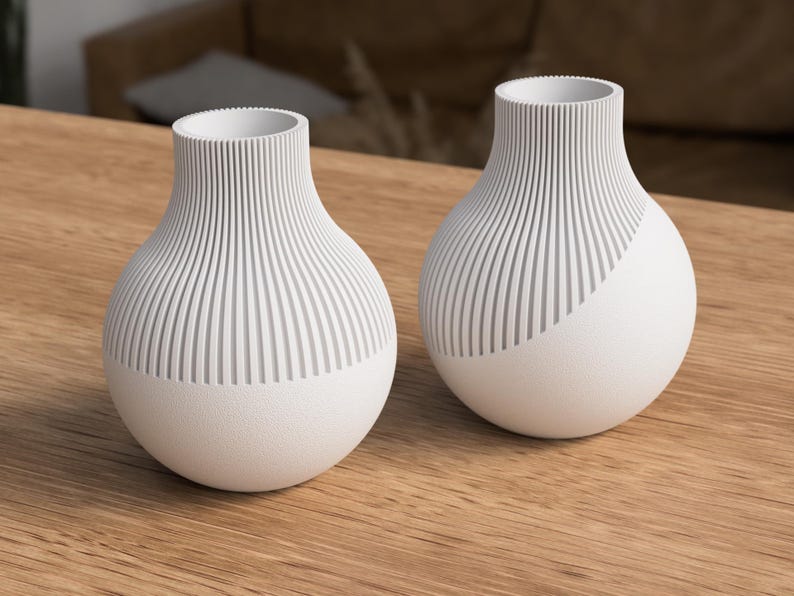May include: Two white ceramic vases with a textured, ribbed design on the upper portion and a smooth, rounded base. One vase has a diagonal design. The vases are displayed on a wooden surface, showcasing a modern, minimalist aesthetic.