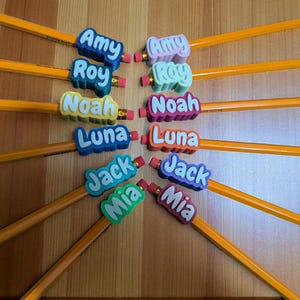 May include: A collection of yellow pencils with colorful name toppers. The toppers feature names like Amy, Roy, Noah, Luna, Jack, and Mia in various colors. Each pencil has a pink eraser.