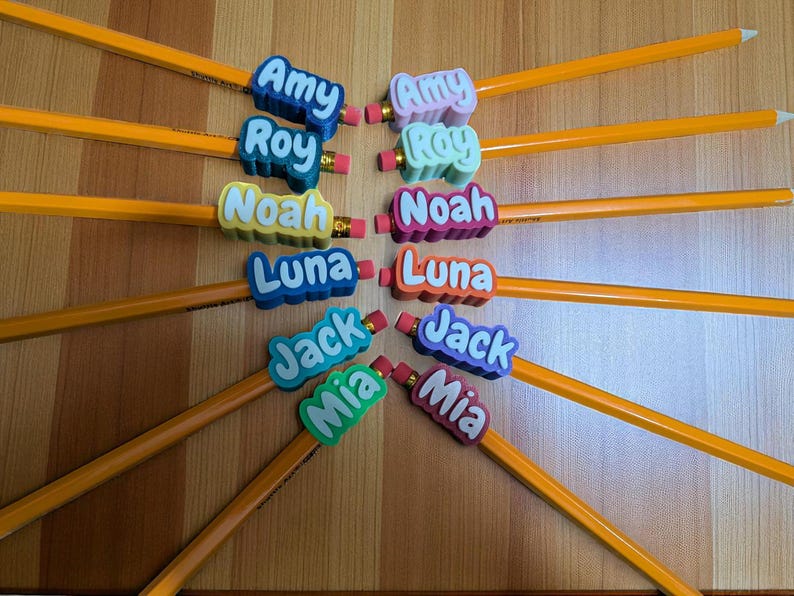 May include: A collection of yellow pencils with colorful name toppers. The toppers feature names like Amy, Roy, Noah, Luna, Jack, and Mia in various colors. Each pencil has a pink eraser.