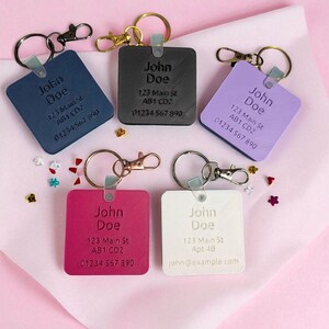 May include: A collection of personalized square keychains in various colors: navy, black, lavender, fuchsia, and white. Each keychain features the name "John Doe", an address, and a phone number. The keychains have metal rings and clasps.
