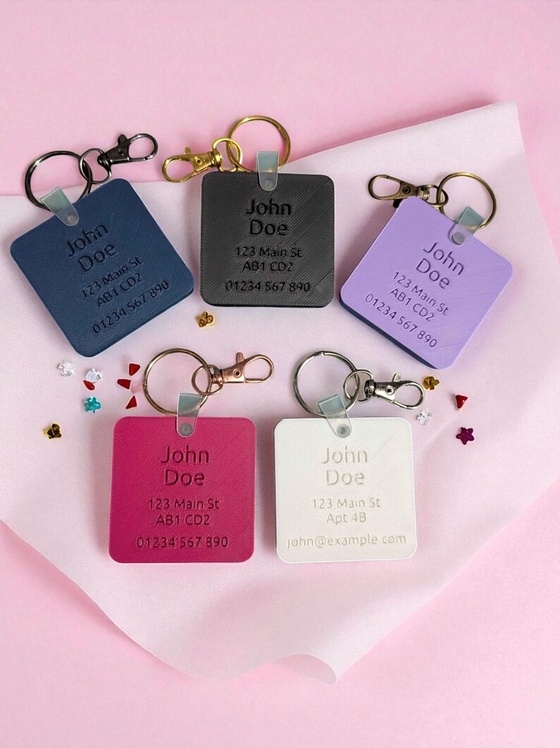 May include: A collection of personalized square keychains in various colors: navy, black, lavender, fuchsia, and white. Each keychain features the name "John Doe", an address, and a phone number. The keychains have metal rings and clasps.