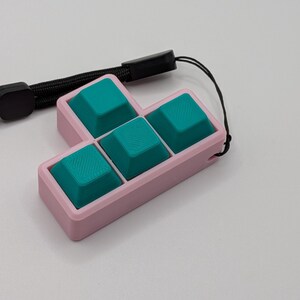 May include: A pink, rectangular keycap holder with four teal keycaps arranged in a Tetris-like shape. A black wrist strap is attached to the holder. The keycaps have a textured surface.