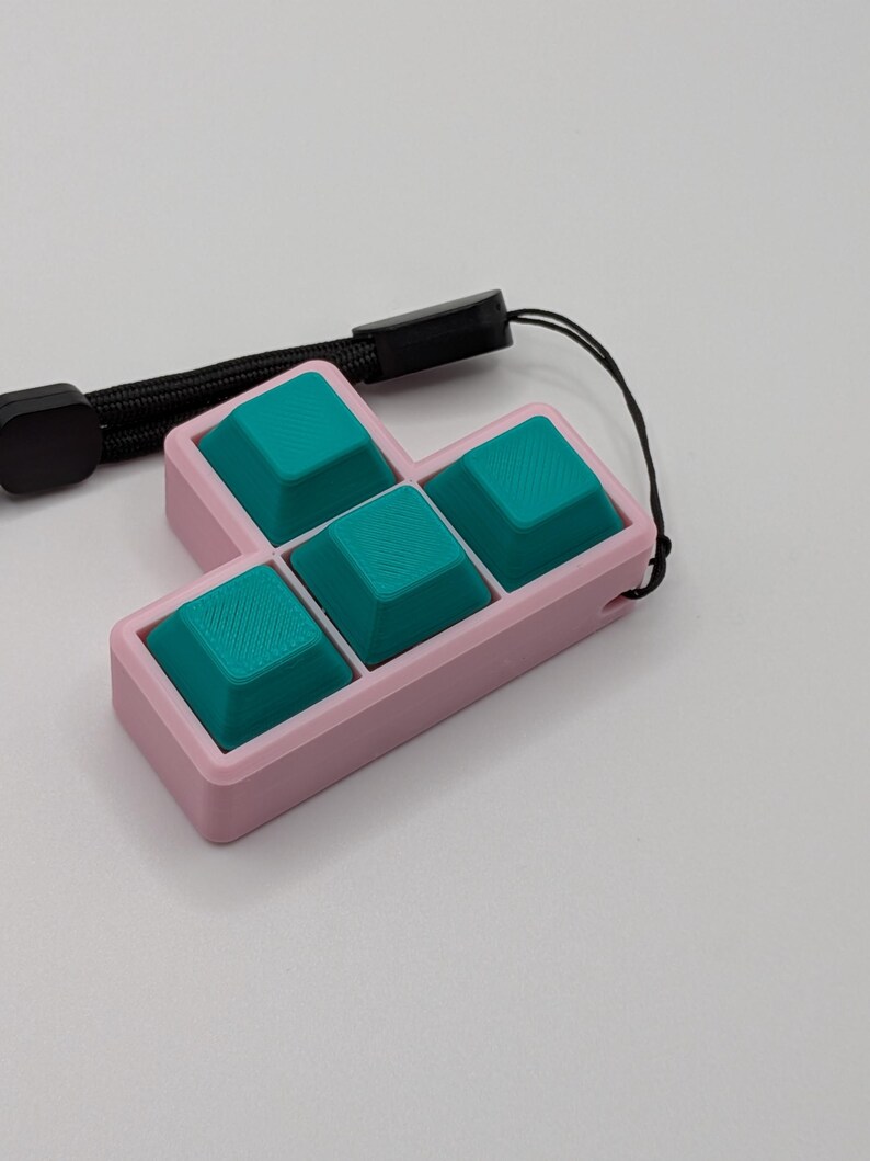 May include: A pink, rectangular keycap holder with four teal keycaps arranged in a Tetris-like shape. A black wrist strap is attached to the holder. The keycaps have a textured surface.