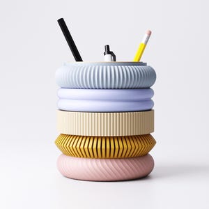 K&ouml;nnte beinhalten: Ein farbenfroher Schreibtisch-Organizer mit gestapeltem Design. Der Organizer besteht aus f&uuml;nf ringf&ouml;rmigen Abschnitten in Pastellfarben: Hellblau, Lavendel, Creme, Gelb und Rosa. Ein schwarzer Stift, ein wei&szlig;er Stift und ein gelber Bleistift sind darin platziert.