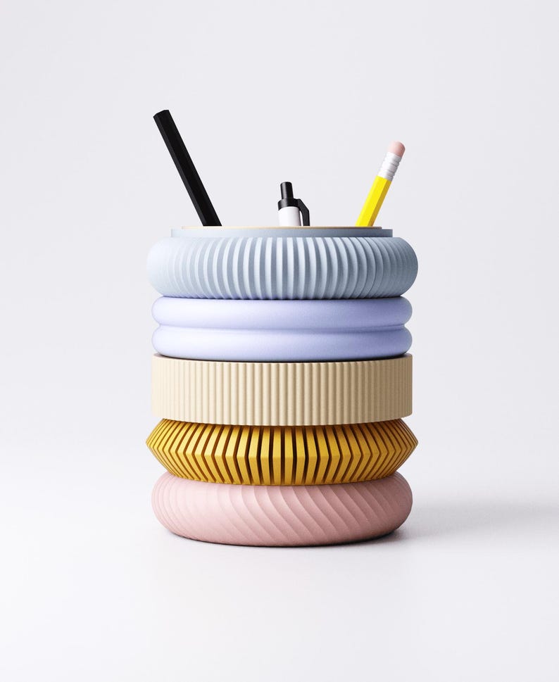 K&ouml;nnte beinhalten: Ein farbenfroher Schreibtisch-Organizer mit gestapeltem Design. Der Organizer besteht aus f&uuml;nf ringf&ouml;rmigen Abschnitten in Pastellfarben: Hellblau, Lavendel, Creme, Gelb und Rosa. Ein schwarzer Stift, ein wei&szlig;er Stift und ein gelber Bleistift sind darin platziert.