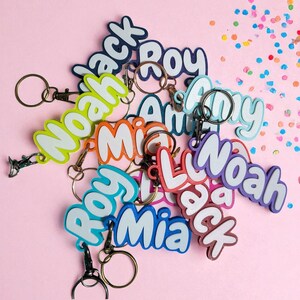 May include: A collection of colorful name keychains with various names like "Noah", "Jack", and "Mia" in a playful font. Each keychain has a metal ring and clasp. The background is a solid pink with scattered confetti.