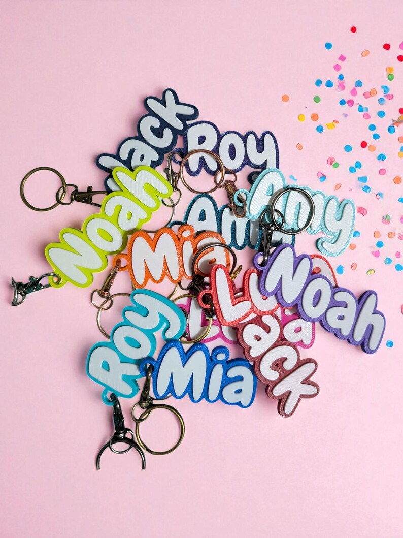 May include: A collection of colorful name keychains with various names like "Noah", "Jack", and "Mia" in a playful font. Each keychain has a metal ring and clasp. The background is a solid pink with scattered confetti.