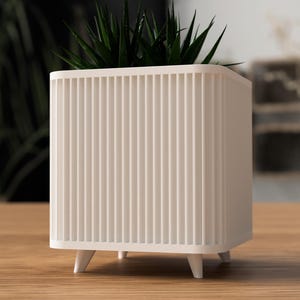 May include: A cream-coloured, square planter with vertical ribbed detailing. The planter has short, angled legs and contains a green succulent plant. The design is modern and minimalist, suitable for indoor or outdoor use.
