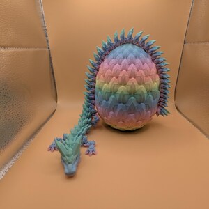 May include: A colorful, 3D-printed dragon egg with a matching dragon. The egg is a gradient of pastel colors, and the dragon is a teal color. The dragon is lying down next to the egg.