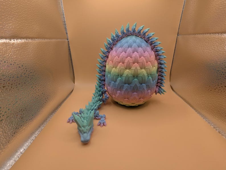 May include: A colorful, 3D-printed dragon egg with a matching dragon. The egg is a gradient of pastel colors, and the dragon is a teal color. The dragon is lying down next to the egg.
