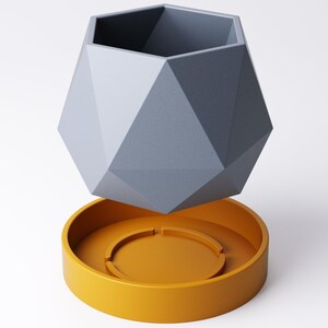 May include: A geometric, gray planter with a faceted design, hovering above a round, mustard-yellow saucer. The planter has a hexagonal opening and is made of a matte material. The saucer has a circular indentation.