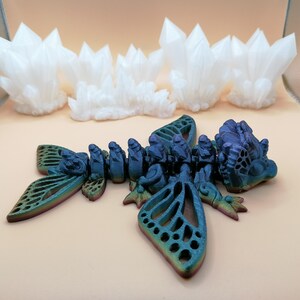May include: A multi-colored, articulated dragon figurine with intricate details, including openwork wings and a segmented body. The dragon is in shades of blue, green, and purple. In the background are several white crystal formations.