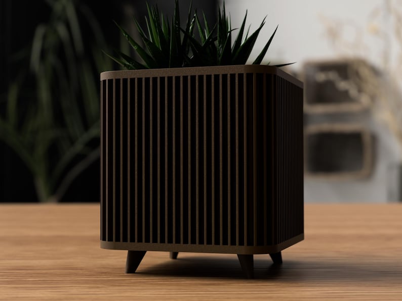 May include: A brown wooden planter with a square shape and vertical slat design. The planter has four small, dark brown legs and holds a green plant with pointed leaves. It rests on a light brown wooden surface.