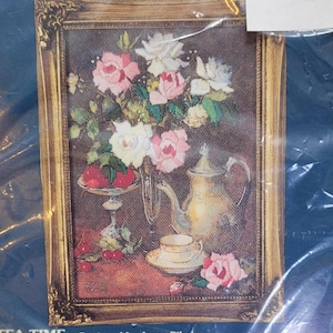 May include: Cross-stitch kit featuring a floral still life with pink and white roses, a teapot, teacup, and fruit. The artwork is framed in a gold-colored ornate frame. The text "TEA TIME" and "Designed by Joyce Pike" are visible. Frame size is 5" x 7".