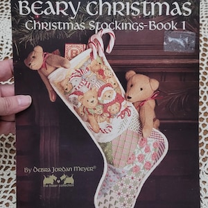 May include: A Leisure Arts craft leaflet titled Beary Christmas Christmas Stockings-Book 1 by Debra Jordan Meyer. The cover features a cross-stitch stocking design with teddy bears, candy canes, and a patchwork pattern, accented by two plush bears.
