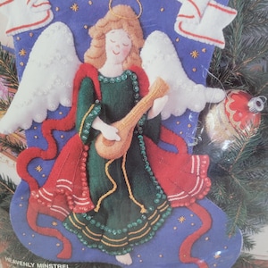 May include: A felt Christmas stocking depicting an angel playing a lute. The angel is dressed in a green gown with red details and white wings. The stocking is blue with gold stars and the text "HEAVENLY MINSTREL".