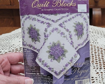 May include: A Dimensions Quilt Blocks stamped cross stitch kit featuring a set of six 15 inch by 15 inch white fabric squares with a purple violet floral pattern. The packaging includes instructions and a thread list for the project.