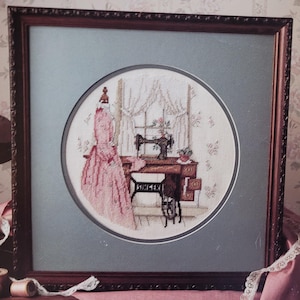 May include: Framed cross-stitch embroidery depicting a vintage sewing room. A pink dress form is positioned near a window with curtains. A Singer sewing machine is on a wooden table. The artwork is framed in a dark brown wooden frame.