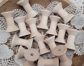 May include: A collection of light-colored wooden spools with a smooth finish, each featuring a central cylindrical core and flared ends, arranged in a scattered pile to showcase their uniform shape and natural grain texture.
