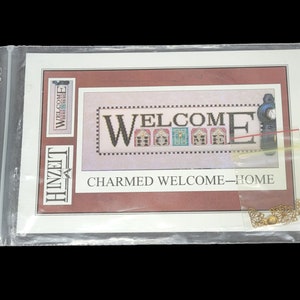 May include: A cross-stitch pattern for a welcome sign with a red, white and blue colour scheme. The pattern features the word "WELCOME" with houses in the letters. The pattern is titled "CHARMED WELCOME HOME" and is packaged in a clear plastic bag.