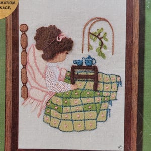 May include: Embroidered artwork in a wooden frame depicts a person seated at a table with a teapot and cups. The person is wearing a pink dress and sitting on a bed. The quilt is green with pink squares. A window with a plant is visible.