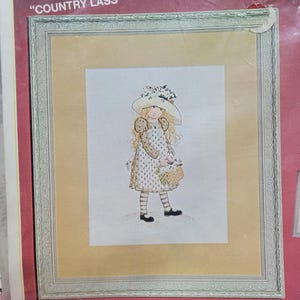 May include: Framed cross-stitch artwork depicting a young girl in a floral dress and hat, carrying a basket of flowers. The artwork is set against a cream background within a decorative silver frame. The text "COUNTRY LASS" is visible at the top.