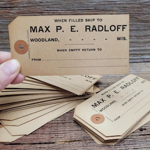 May include: A stack of rectangular tan shipping tags with reinforced circular holes. Each tag features black printed text reading WHEN FILLED SHIP TO MAX P. E. RADLOFF WOODLAND, WIS. and WHEN EMPTY RETURN TO FROM with blank lines for writing.