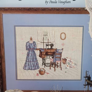 May include: Cross-stitch artwork showing a sewing room. A blue dress form stands near a sewing machine and chair. The artwork is framed with a light blue background and a wooden frame.