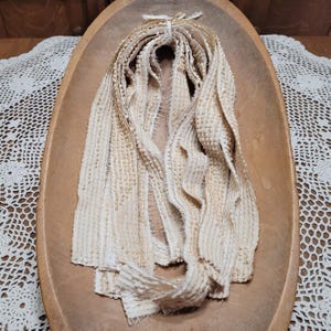 May include: Cream-colored, textured fabric strips tied together, displayed in a wooden bowl. The strips have a woven appearance with a slightly frayed edge. The bowl is oval-shaped and sits on a white lace tablecloth.
