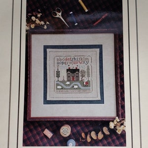 May include: A framed cross-stitch pattern featuring a house with a red door and white trim, surrounded by trees and sheep. The text "The Brown Ranch - 1915 - A.M. Scott County - CA" is stitched below the house. The image is surrounded by a brown frame and a plaid fabric background.