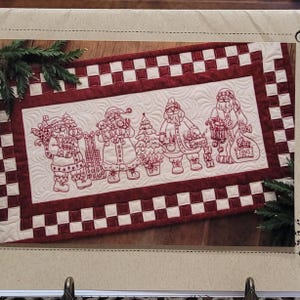 May include: A rectangular Christmas table runner with embroidered Santa Claus figures in red thread on a cream background. The runner has a red and white checkered border and a quilted design. The Santas are carrying gifts and a Christmas tree.