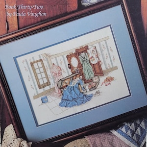 May include: Cross-stitch picture in a dark wood frame. The artwork shows a room with a wardrobe, dresses, and a dress form. A blue dress is in the foreground. The text "Book Thirty-Two by Paula Vaughan" is in the upper left corner.