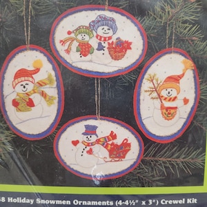 May include: Four oval-shaped holiday snowman ornaments. Each ornament features a snowman with a hat, scarf, and other accessories. The ornaments are approximately 11.4 cm by 7.6 cm.