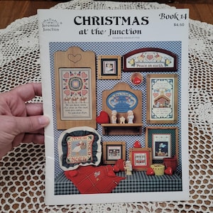 May include: A Jeremiah Junction Christmas at the Junction Book 14 counted cross stitch pattern booklet. The cover features various finished cross stitch projects including framed designs, a small shelf with figurines, and a red plaid fabric piece.