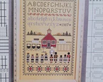 May include: A framed cross-stitch sampler on a light beige fabric featuring an alphabet in uppercase and lowercase letters, the year 1987, a red schoolhouse, a teacher, and students standing behind a white fence, bordered by geometric star patterns.