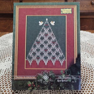 May include: A cross-stitch pattern book titled "Oh Christmas Tree!" featuring a detailed Christmas tree design with red and white accents. The cover has a green border and a red inner frame, with the text "CROSS 'N PATCH" and the designer's name.