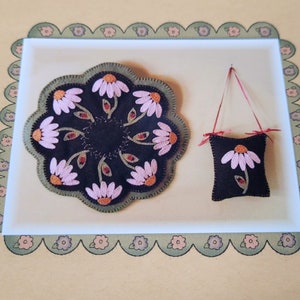 May include: A set of two felt crafts featuring a daisy design. The larger craft is a round rug with a black centre and pink daisy flowers. The smaller craft is a black cushion with a pink daisy flower. Both crafts have green stems and leaves.