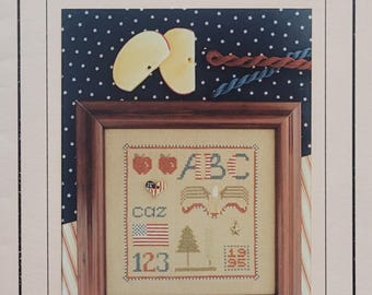 May include: A framed cross-stitch sampler titled The Drawn Thread Charted Needlework Designs. The sampler features red apples, the letters ABC, an eagle, the American flag, a tree, the numbers 123, and the year 1995 on a tan fabric background.