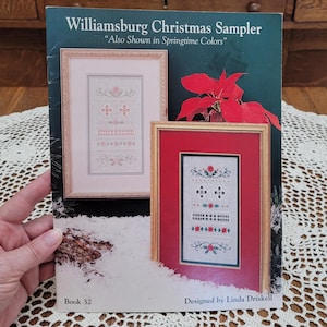 May include: A craft pattern book titled Williamsburg Christmas Sampler, Also Shown in Springtime Colors, Book 32, Designed by Linda Driskell. The cover displays two framed cross-stitch samplers, one in soft pink tones and one in red and green.