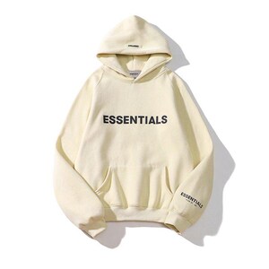 May include: Cream-colored hoodie with the word "ESSENTIALS" printed in black across the chest. The hoodie has a hood, a front pocket, and the word "ESSENTIALS" printed on the left sleeve. A casual, comfortable garment.