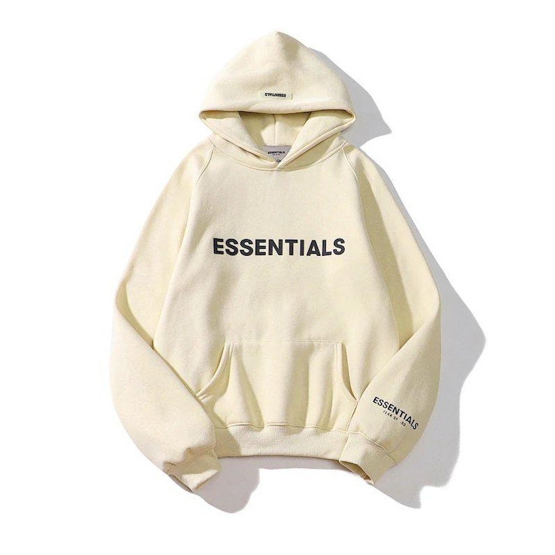 May include: Cream-colored hoodie with the word "ESSENTIALS" printed in black across the chest. The hoodie has a hood, a front pocket, and the word "ESSENTIALS" printed on the left sleeve. A casual, comfortable garment.