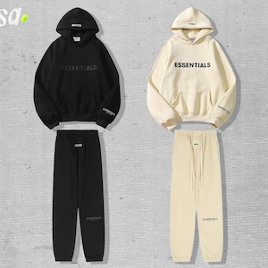 May include: Two matching tracksuits, one black and one cream-colored, each consisting of a hooded sweatshirt and sweatpants. The word "ESSENTIALS" is printed on the front of the sweatshirts. The tracksuits are displayed on a light gray background.