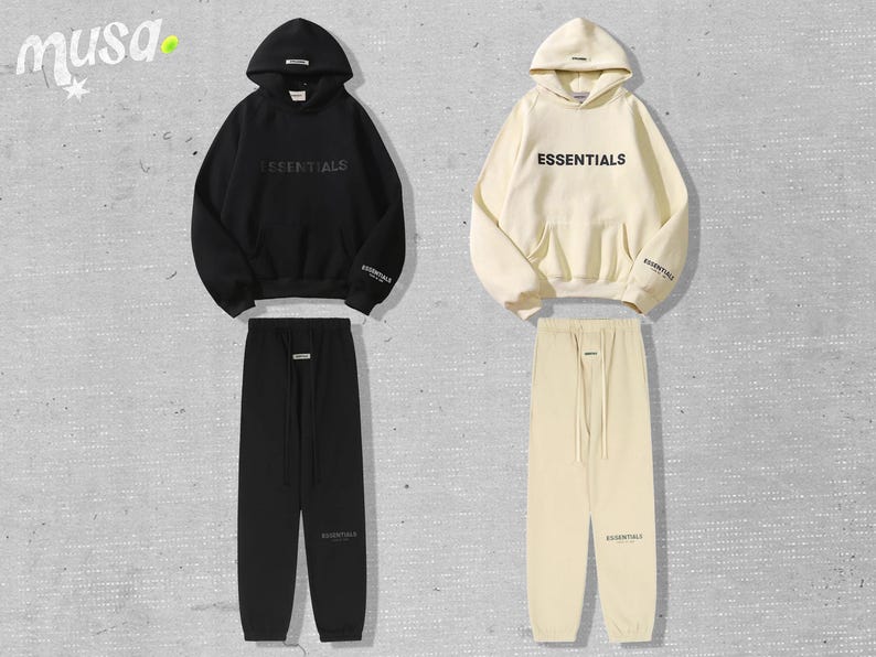 May include: Two matching tracksuits, one black and one cream-colored, each consisting of a hooded sweatshirt and sweatpants. The word "ESSENTIALS" is printed on the front of the sweatshirts. The tracksuits are displayed on a light gray background.