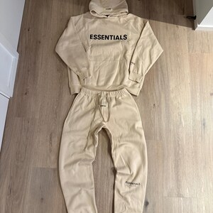 May include: A beige hoodie and matching sweatpants set. The word "ESSENTIALS" is printed in black on the front of the hoodie and on the left leg of the sweatpants. The hoodie has a drawstring hood and a front pocket.